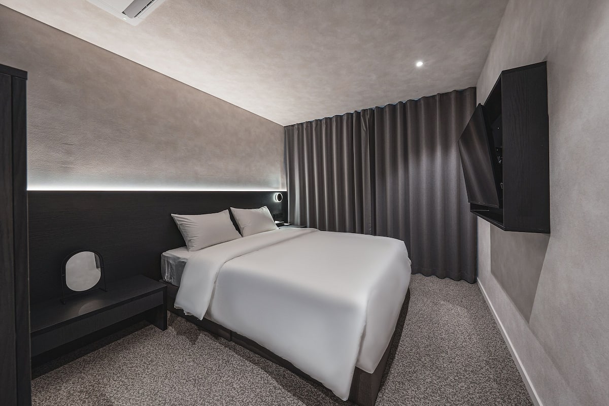 A modern hotel room with a large bed, a bedside table, a wall-mounted TV, and a ceiling light.