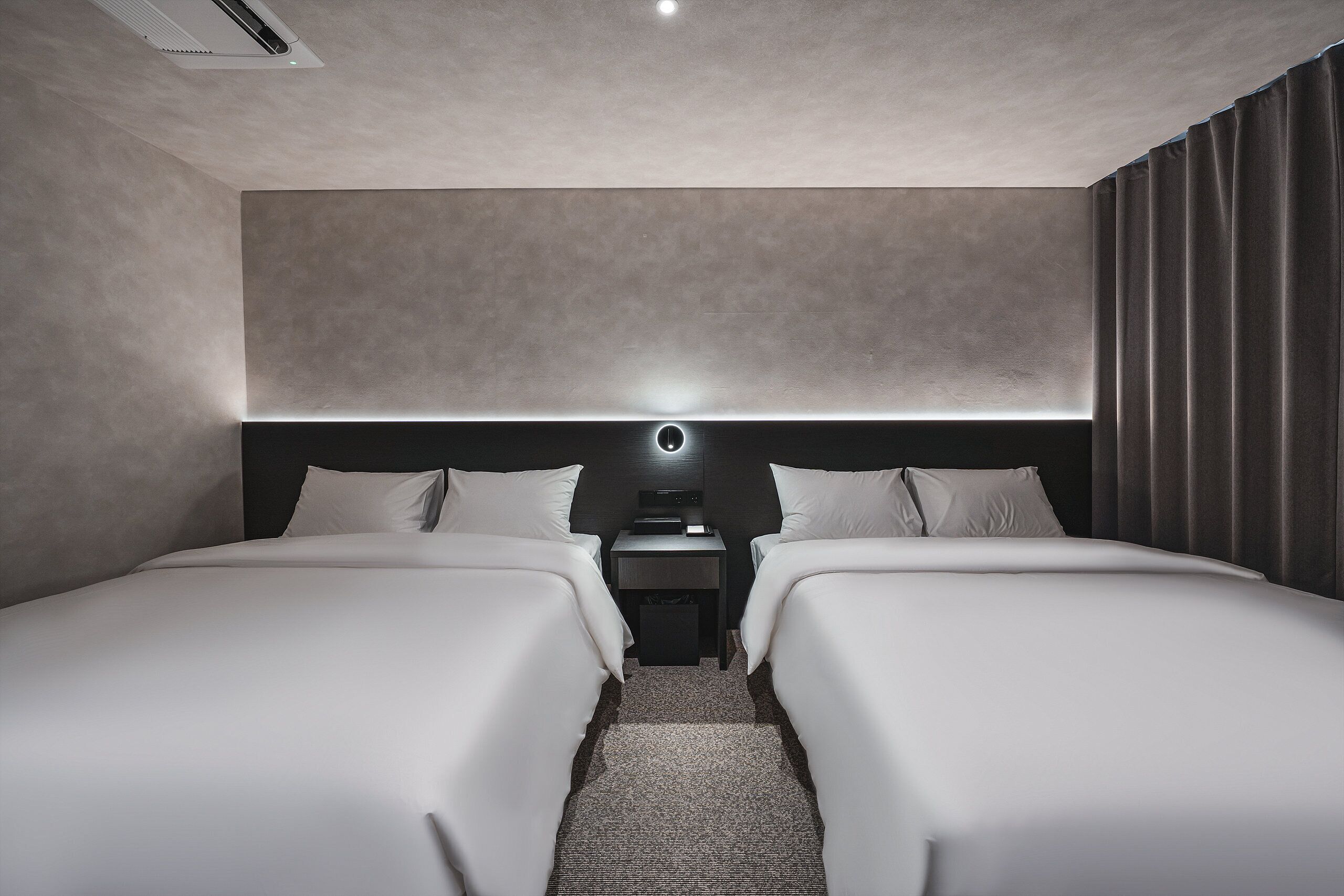 Una habitación de hotel moderna con dos camas, una mesita de noche y un cabecero con iluminación integrada.