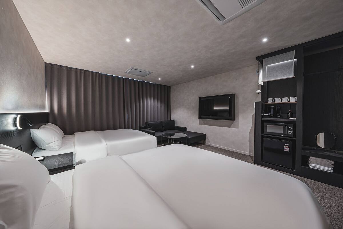 A modern hotel room with two beds, a sofa, a small table, a TV, and a minibar.