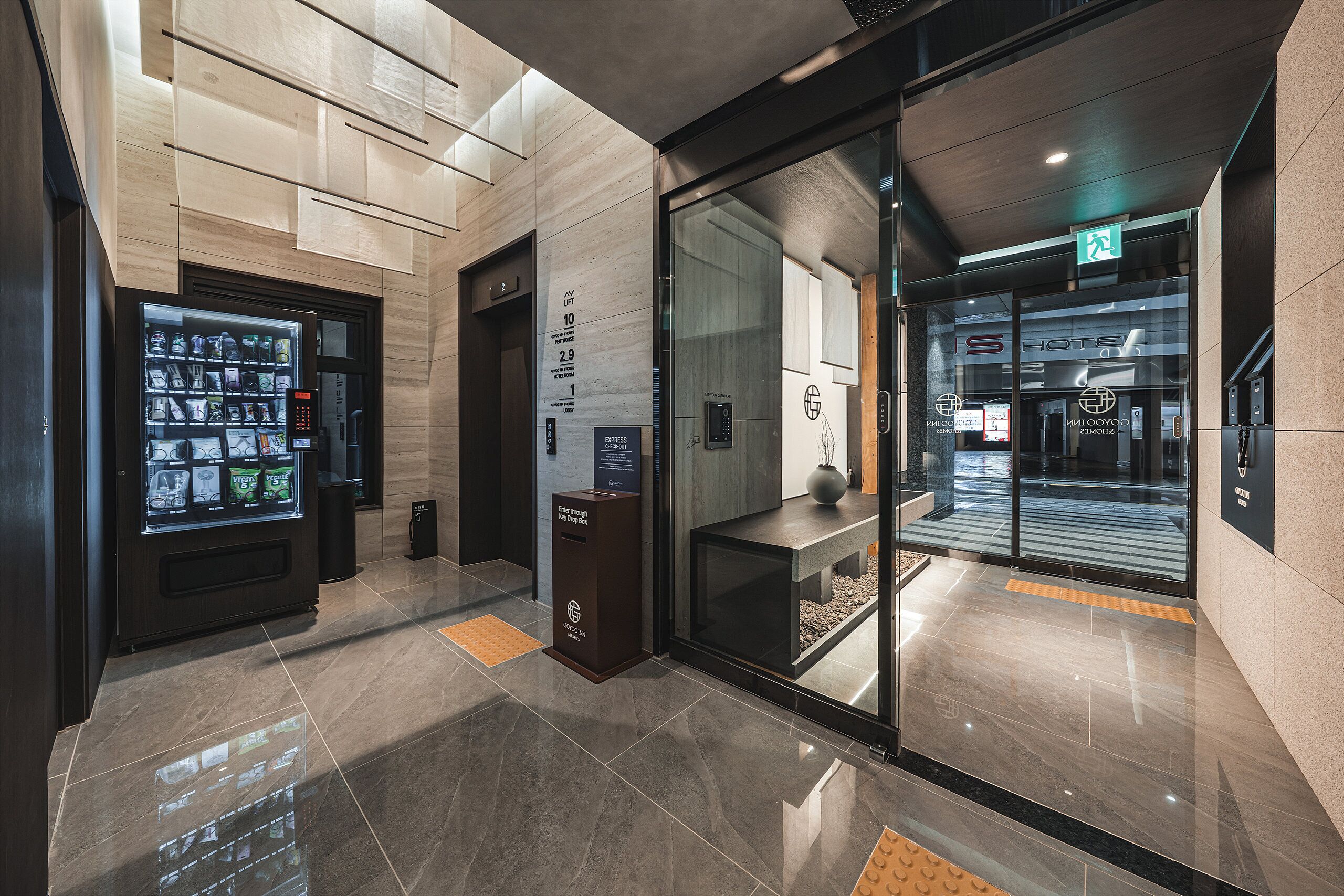Un moderno vestíbulo de hotel con una máquina expendedora, una recepción y una puerta de vidrio que da al ascensor.