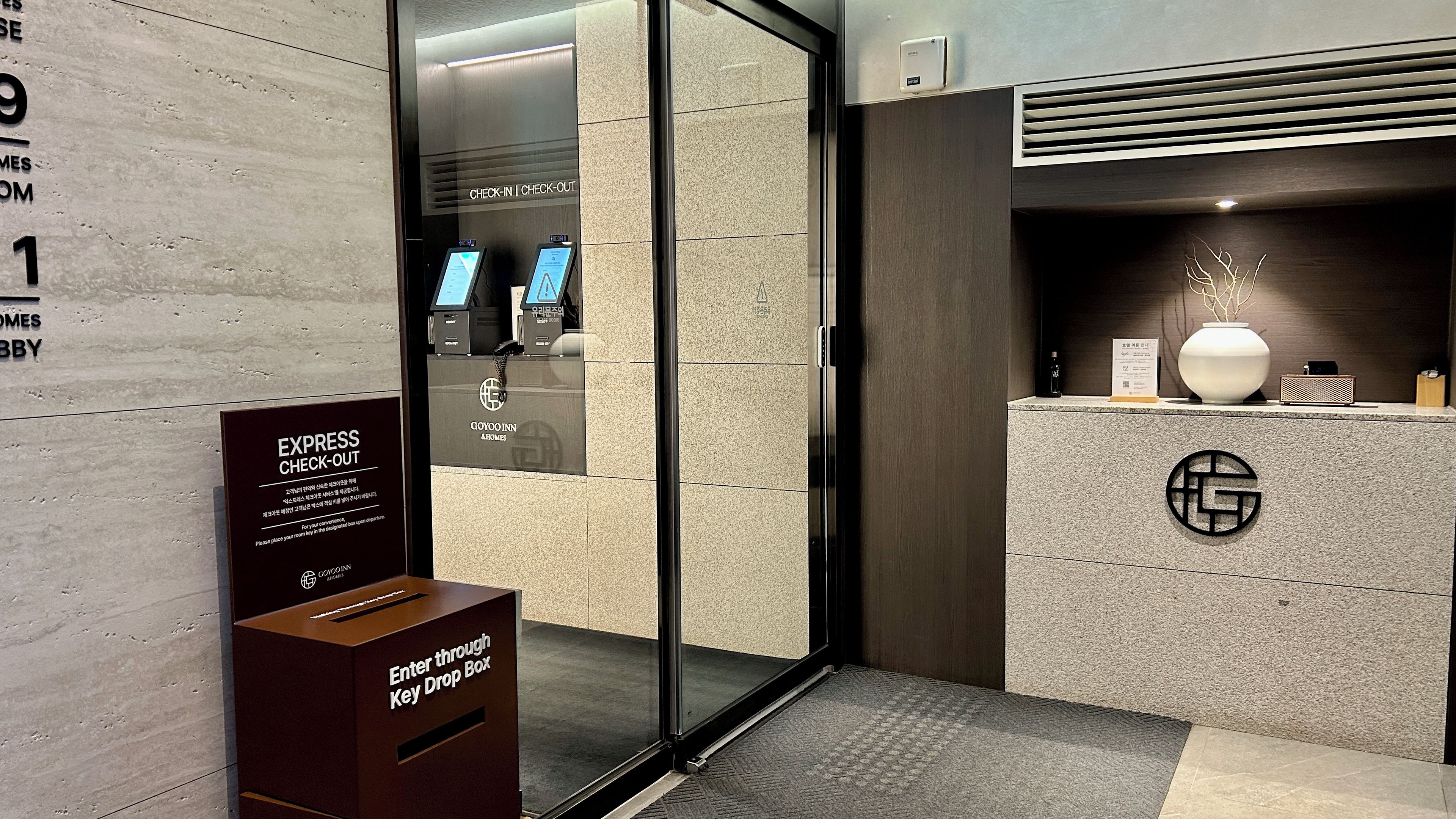 A hotel entrance with a glass door, a key drop box, and a reception desk with a logo.