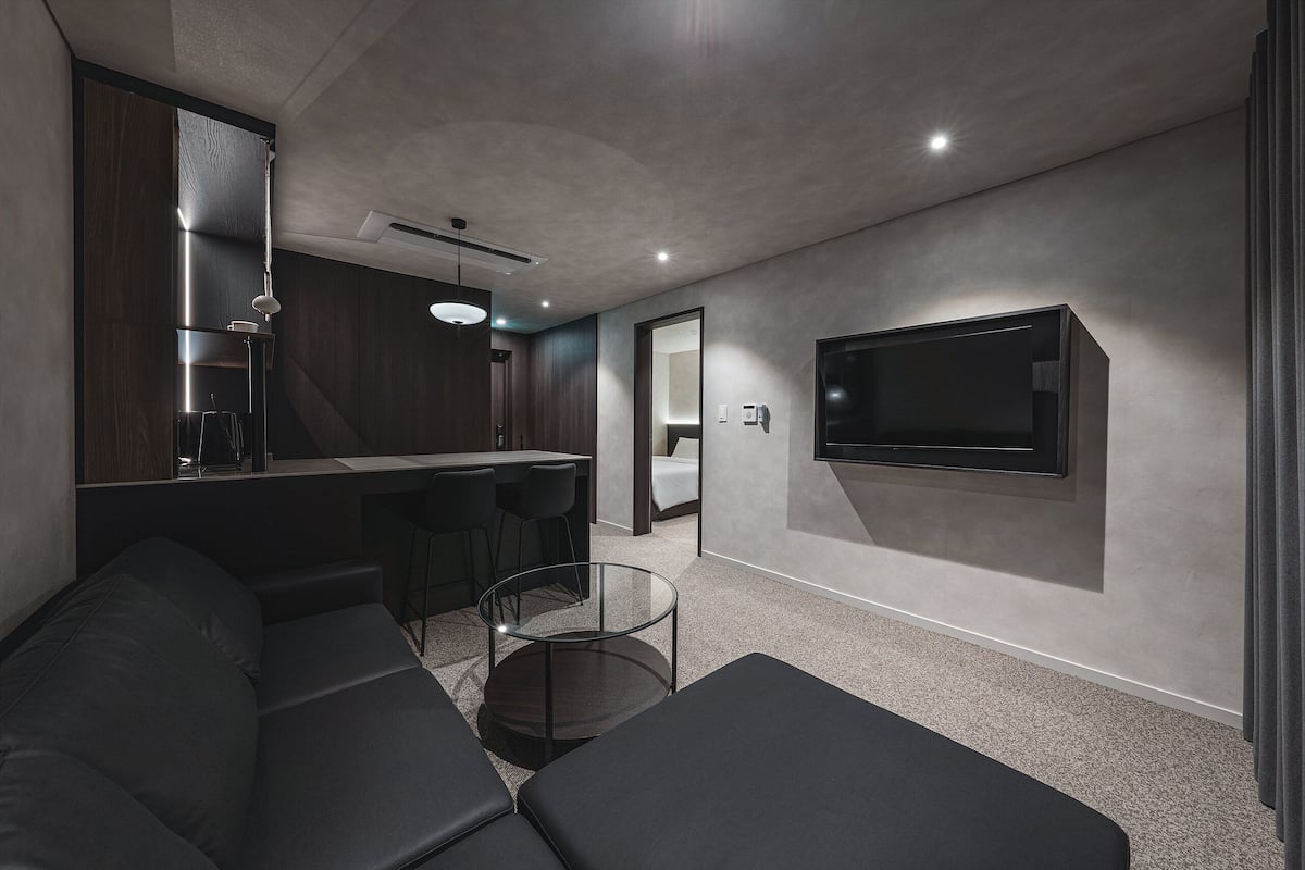A modern living room with a sectional sofa, a glass coffee table, a flat-screen TV mounted on the wall, and a kitchen area with dark wood cabinets and a pendant light.