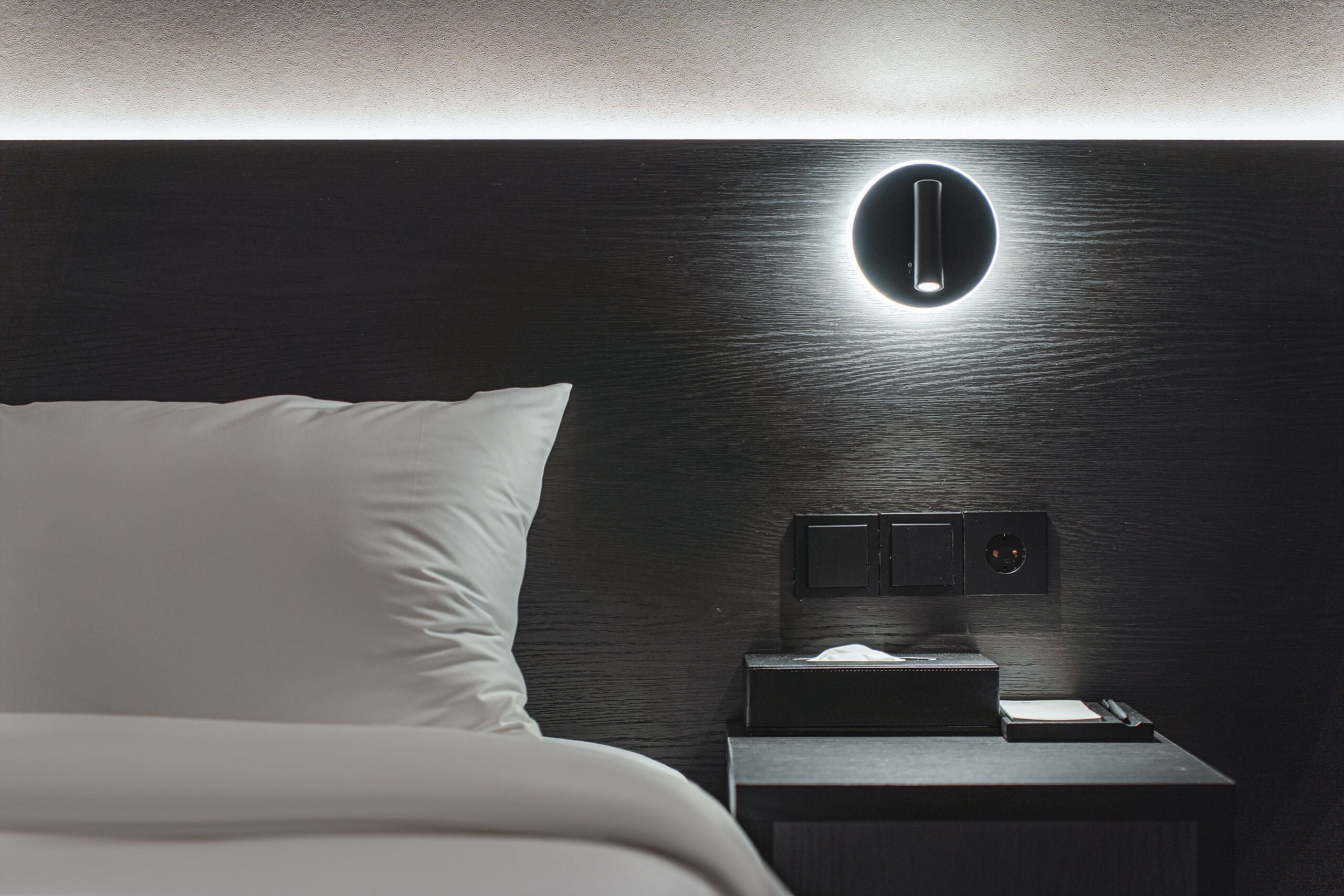 Un lit bien fait, avec du linge de lit blanc, une table de chevet avec une lampe et un luminaire fixé au mur.