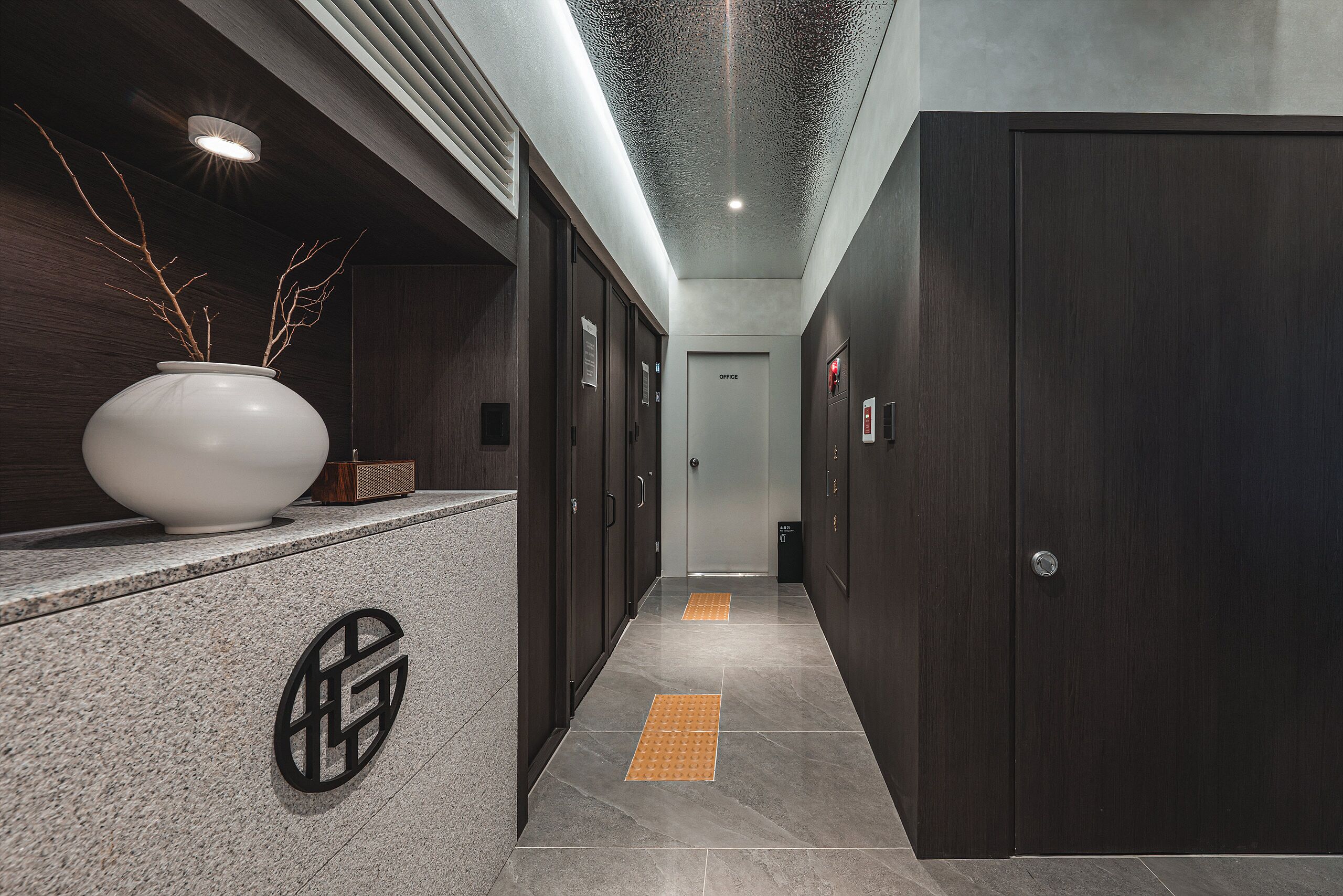 A modern hallway with a reception desk, dark wood doors, and a tiled floor with tactile paving.
