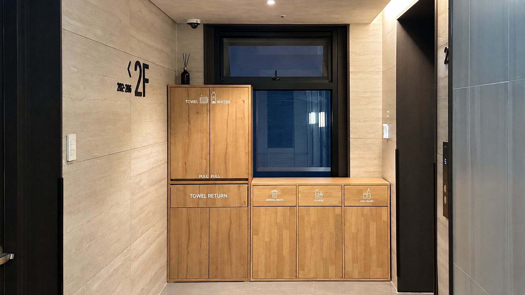 Un baño moderno con un mueble de madera para guardar toallas y artículos de aseo, un cesto para toallas usadas y cubos de basura para residuos generales, plásticos y vidrio.