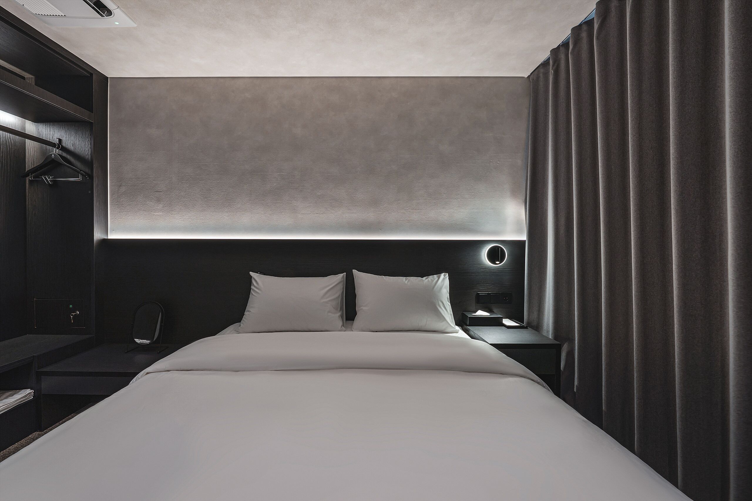 A modern hotel room with a large bed, a bedside table, a nightstand, a wardrobe, and a window with curtains.