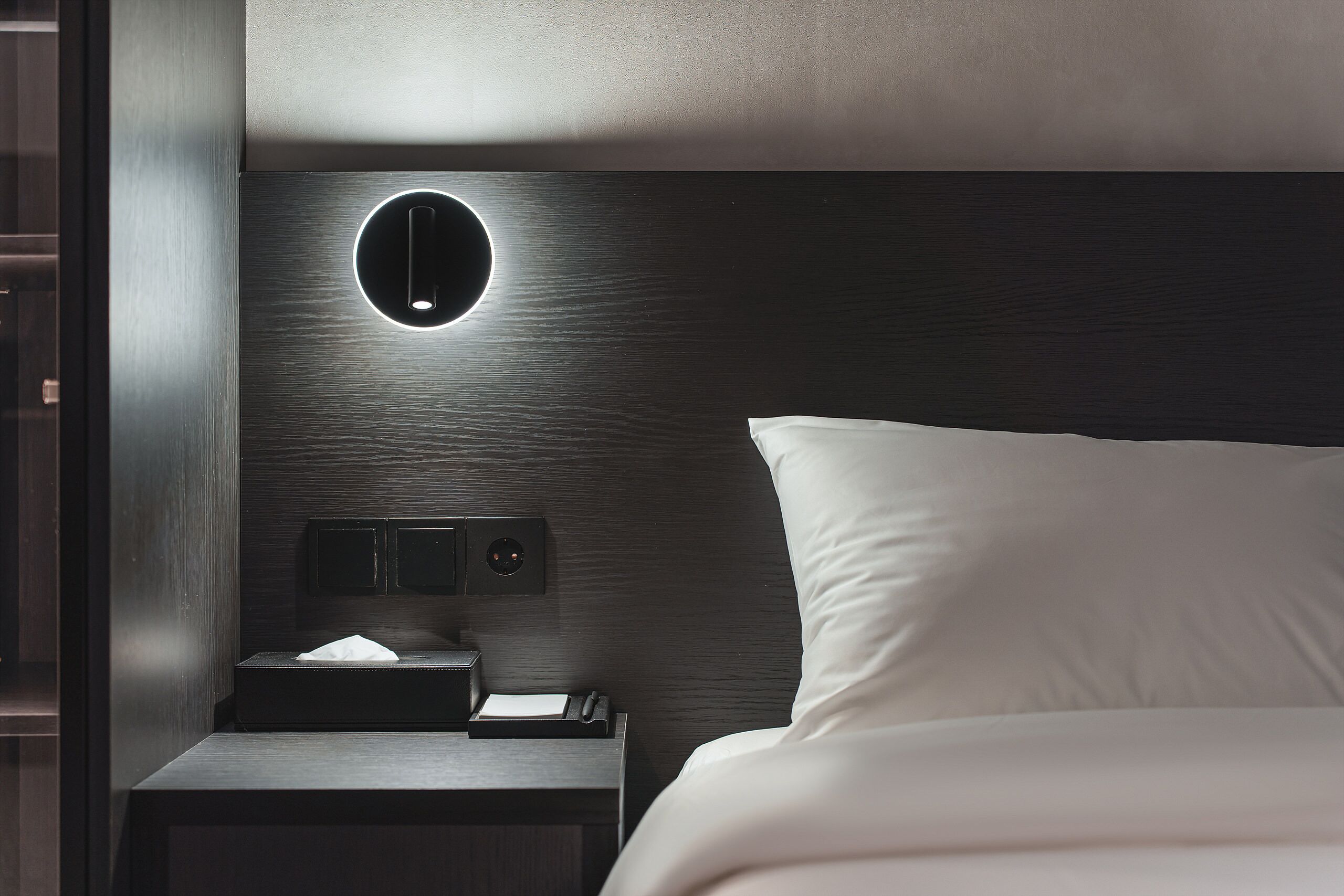 Una habitación de hotel moderna con un cabecero de madera oscura, una mesita de noche con lámpara y una cama bien tendida.