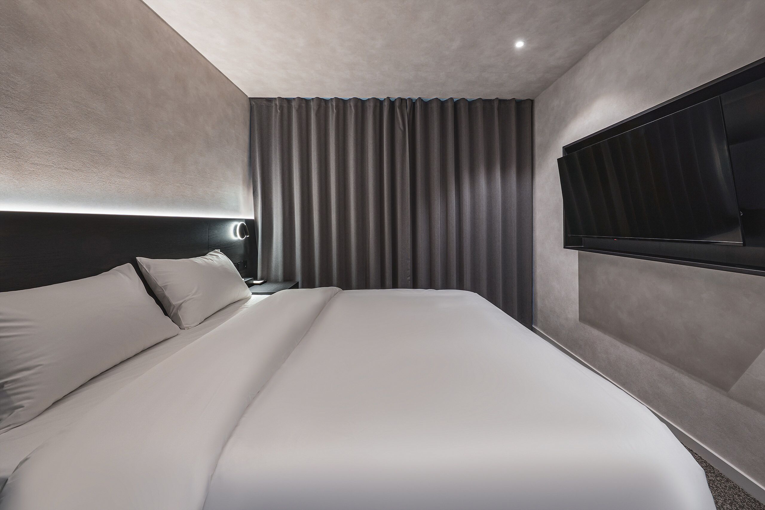 A modern hotel room with a large bed, a flat-screen TV mounted on the wall, and a nightstand with a lamp.