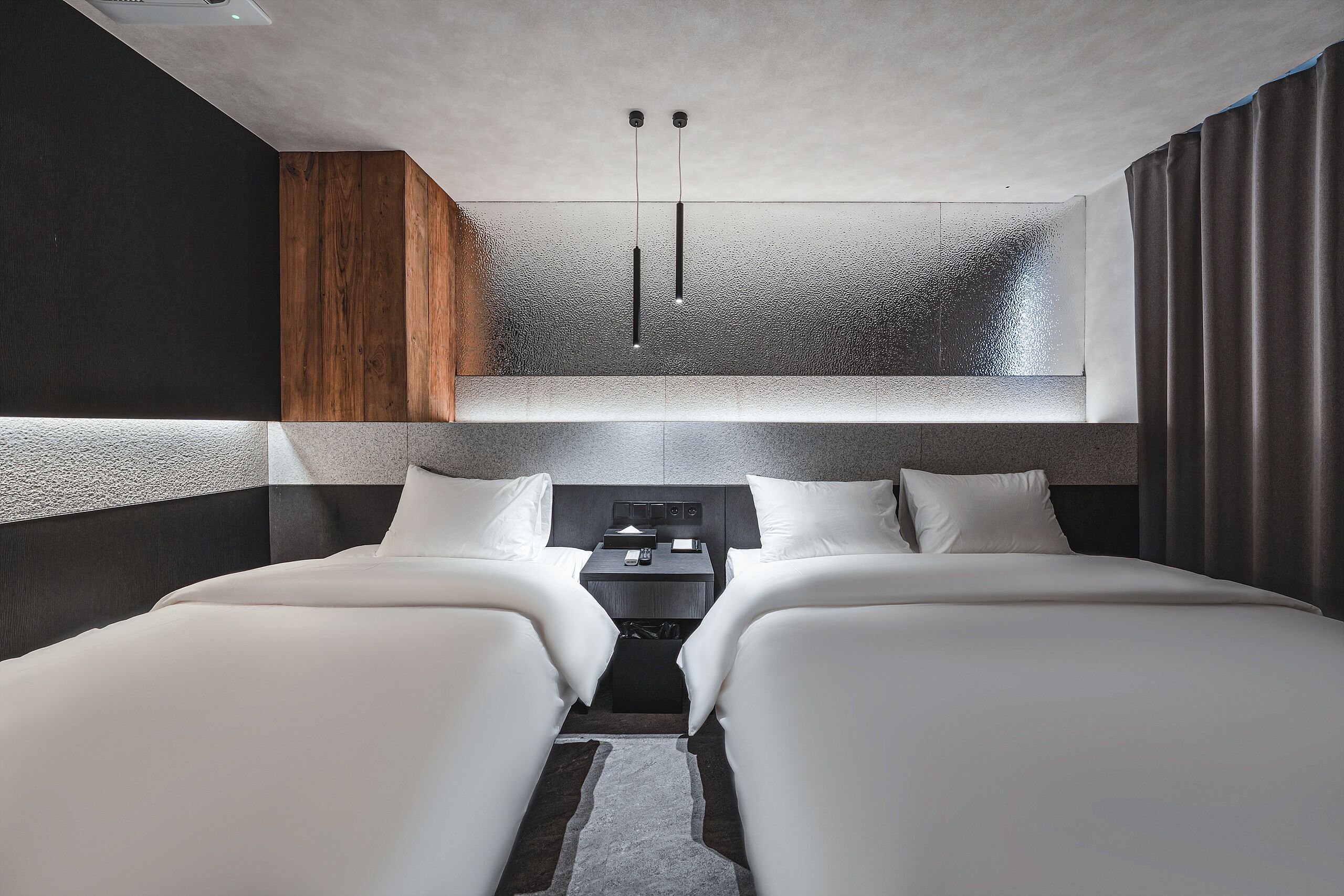 Una habitación de hotel moderna con dos camas, una pared decorativa de madera, una mesita con un control remoto y un aplique de techo.