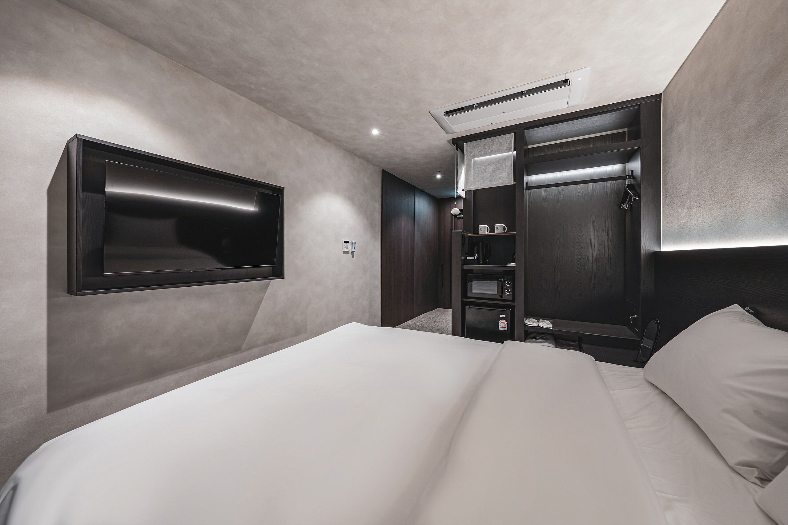 A modern hotel room with a large bed, built-in TV, wardrobe, and a small desk area.