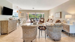 A spacious living room with a large TV, wicker furniture, a sofa set, and a dining area with chairs.