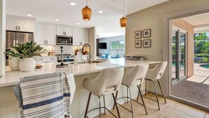 A modern kitchen with white cabinets, a central island with bar stools, a stainless steel refrigerator, and a large window overlooking a pool.