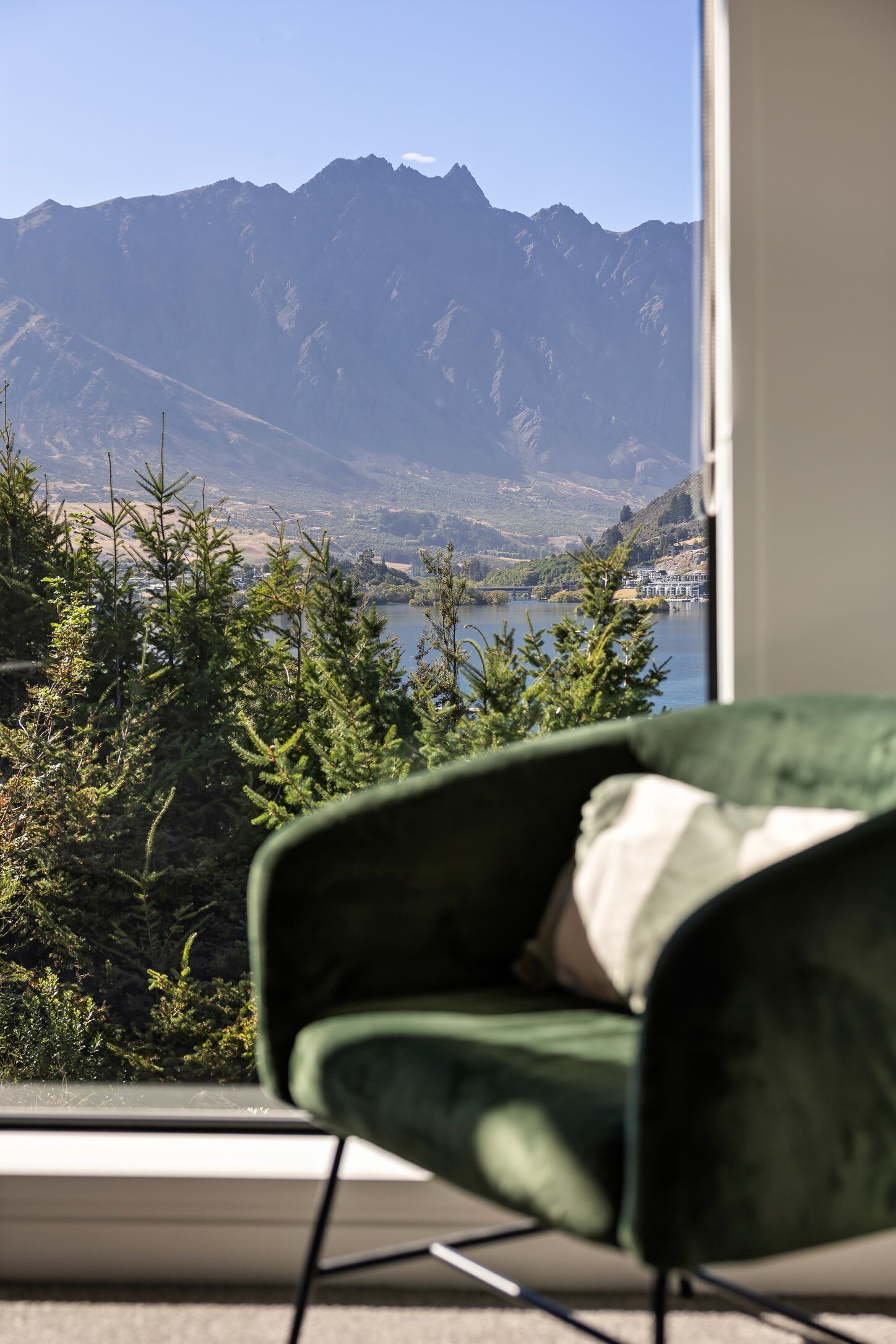 A view from a window with a green chair, overlooking a lake and mountains.