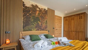 Une chambre à coucher avec un lit, une tête de lit en bois, une table de chevet, une lampe fixée au mur et une grande fenêtre donnant sur la forêt.