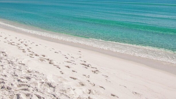 Ein Strand mit Fußabdrücken, die zum Wasser führen, ein klarer Himmel und ruhige Wellen.