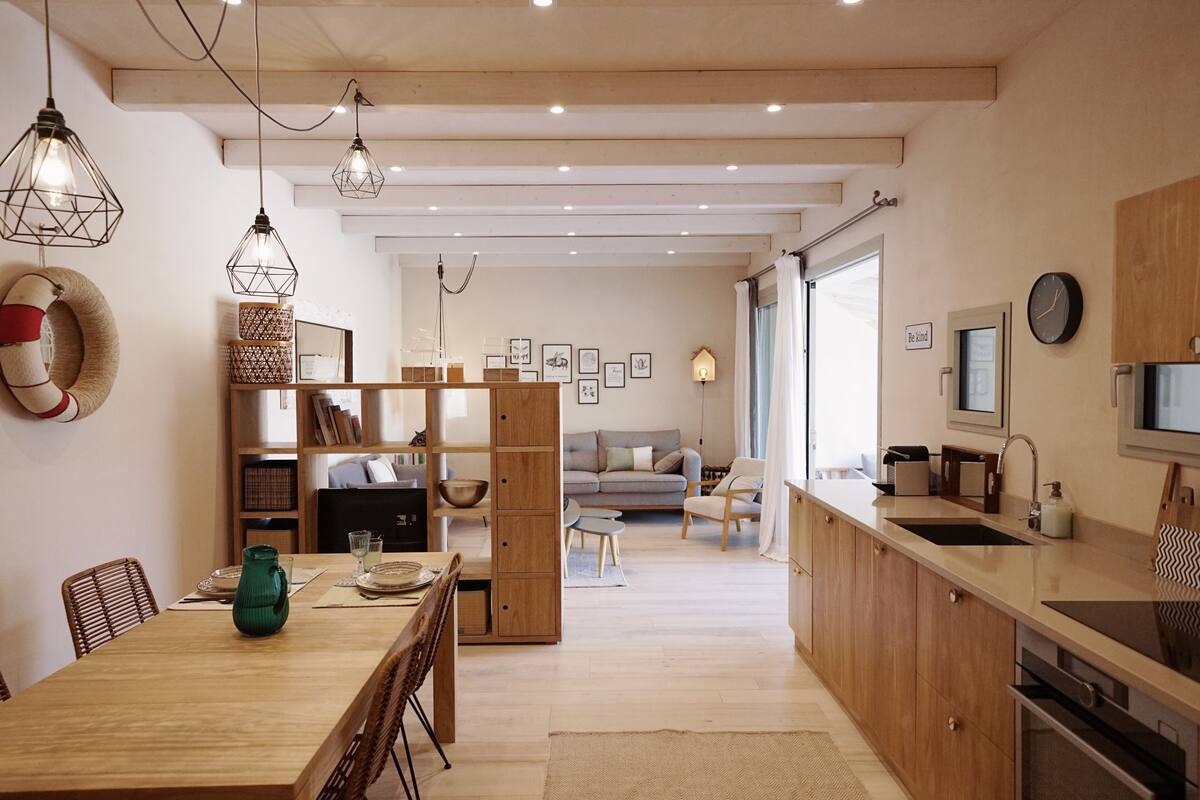 A modern kitchen with wooden cabinets, a dining area with a table and chairs, and a living area with a sofa and a TV.