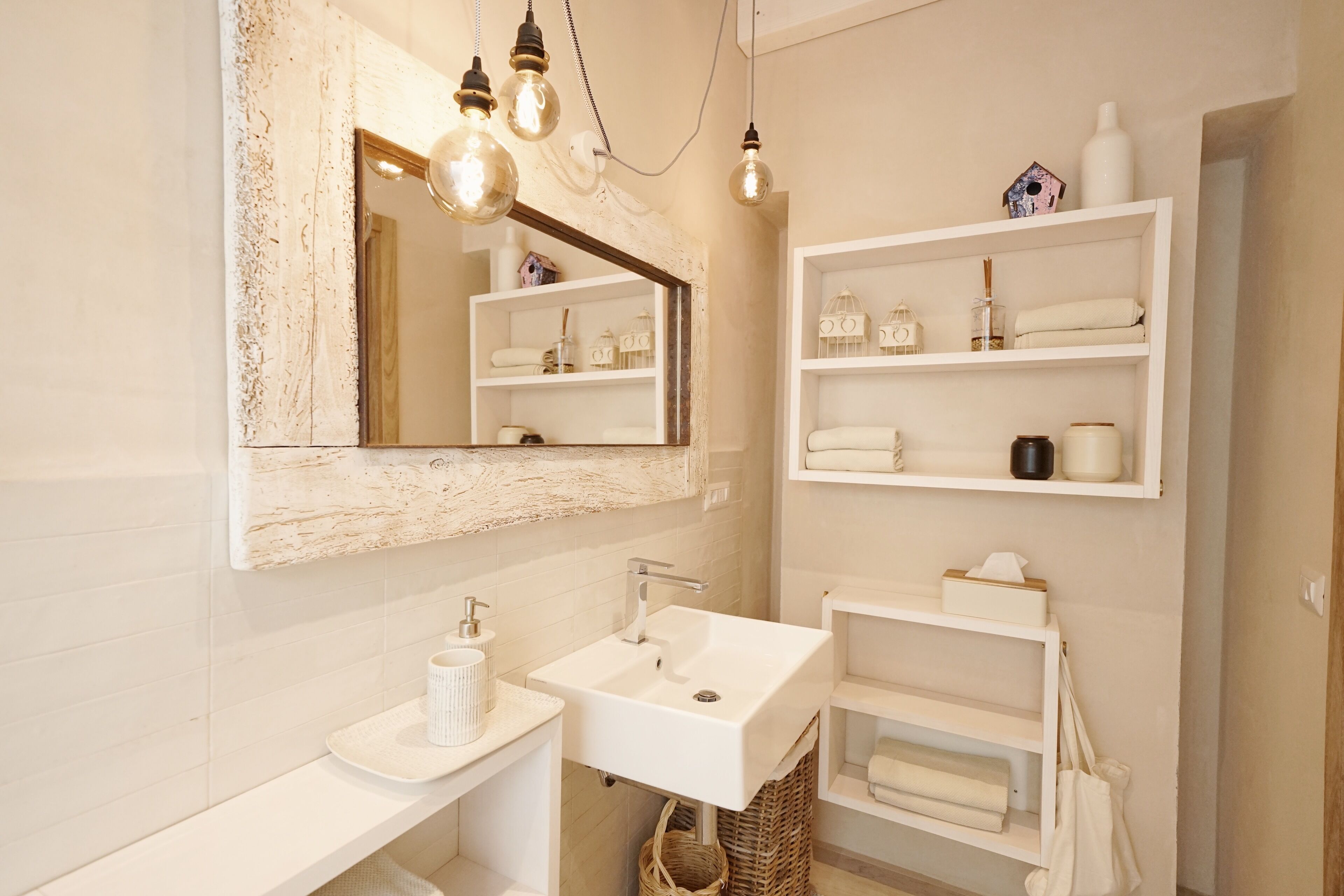 Un baño moderno con lavabo blanco, espejo y estantes con artículos de aseo.