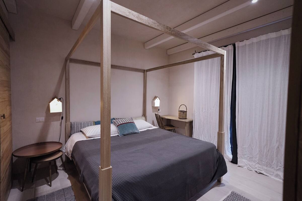 A four-poster bed with a dark bedspread, a bedside table with a lamp, and a desk with a chair and a basket.