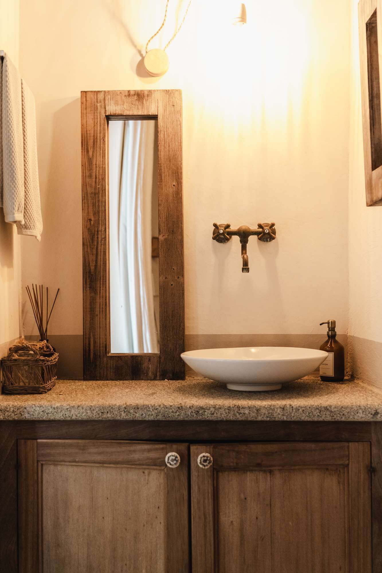 Un baño con tocador de madera, lavamanos blanco, toallero, espejo y una lámpara de pared.