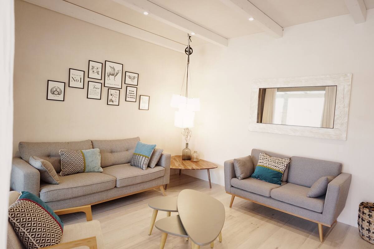 A living room with two grey sofas, a small wooden table, and a floor lamp. There are framed pictures on the wall and a mirror above the sofa.