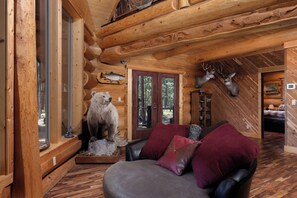 A log cabin interior with a bear and deer mounted on the walls, a leather couch with red cushions, and a wooden floor.