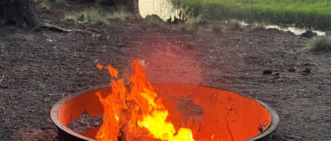 Una fogata en un brasero metálico rodeada de un paisaje natural con árboles y una masa de agua al fondo.