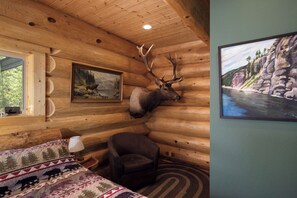 A log cabin room with a mounted elk head, a bed with a patterned quilt, a chair, and a painting of a forest scene.