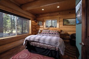 A log cabin bedroom with a large bed, wooden walls, and a window overlooking a forest.