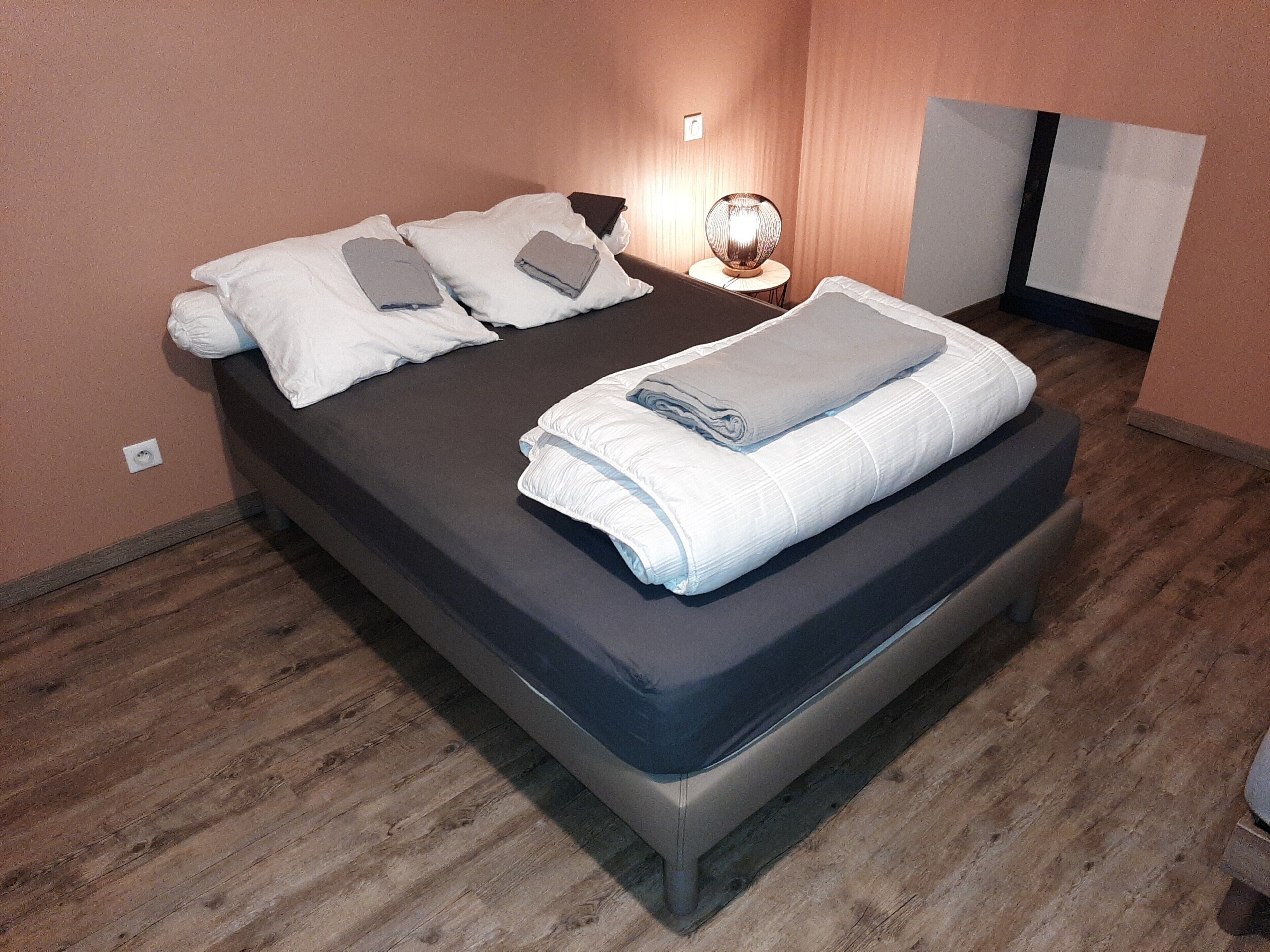 Cama bem arrumada com almofadas e um cobertor, uma luminária de cabeceira e piso de madeira.