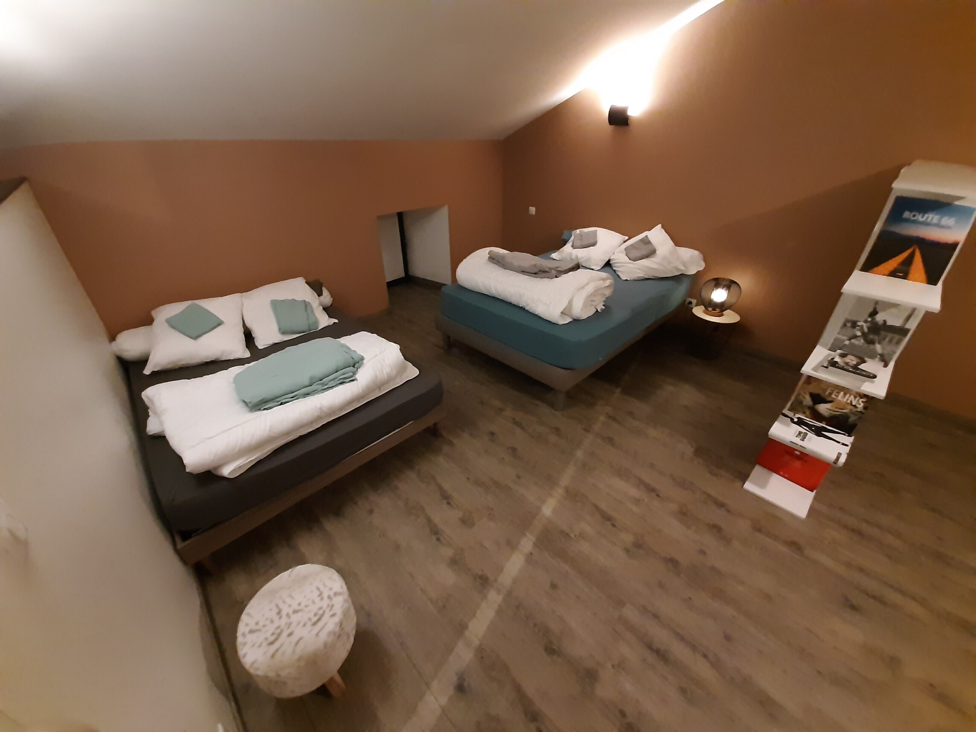 Ein Hotelzimmer mit zwei Betten, einem kleinen Hocker, einem Wandregal mit Broschüren und einer Deckenleuchte.