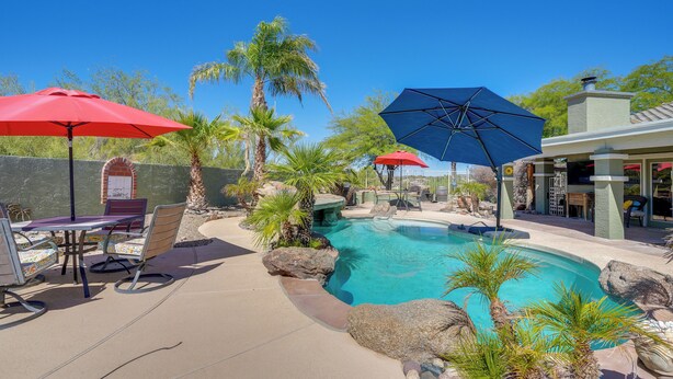 A pool area with a clear blue pool, surrounded by rocks and palm trees. There are outdoor seating areas with umbrellas and a patio with a table and chairs. - Backyard Oasis & Hot Tub: Mtn-view Wickenburg Home (Wickenburg)