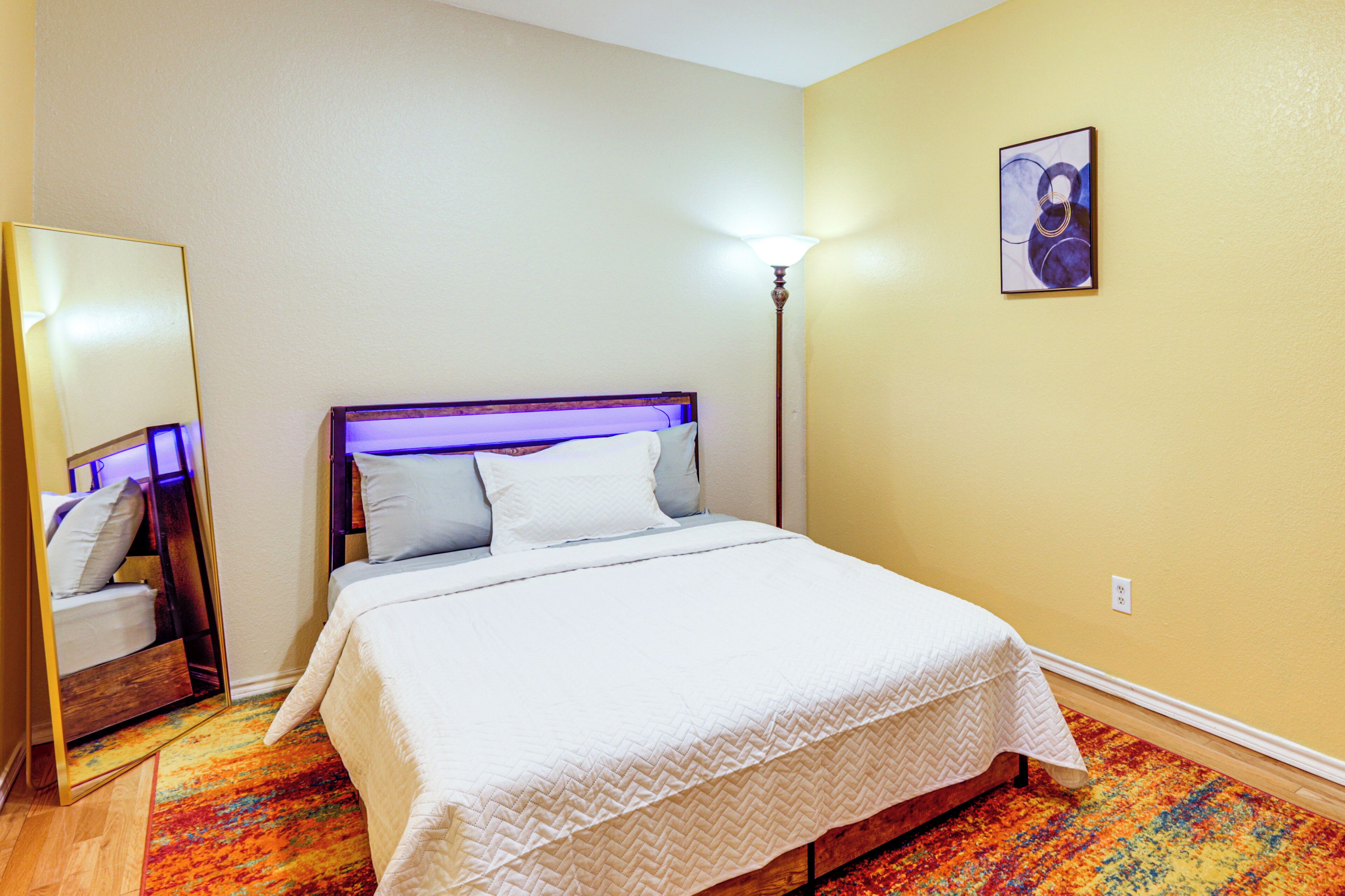 A neatly made bed with a white quilt, a colorful rug, and a mirror reflecting part of the room.