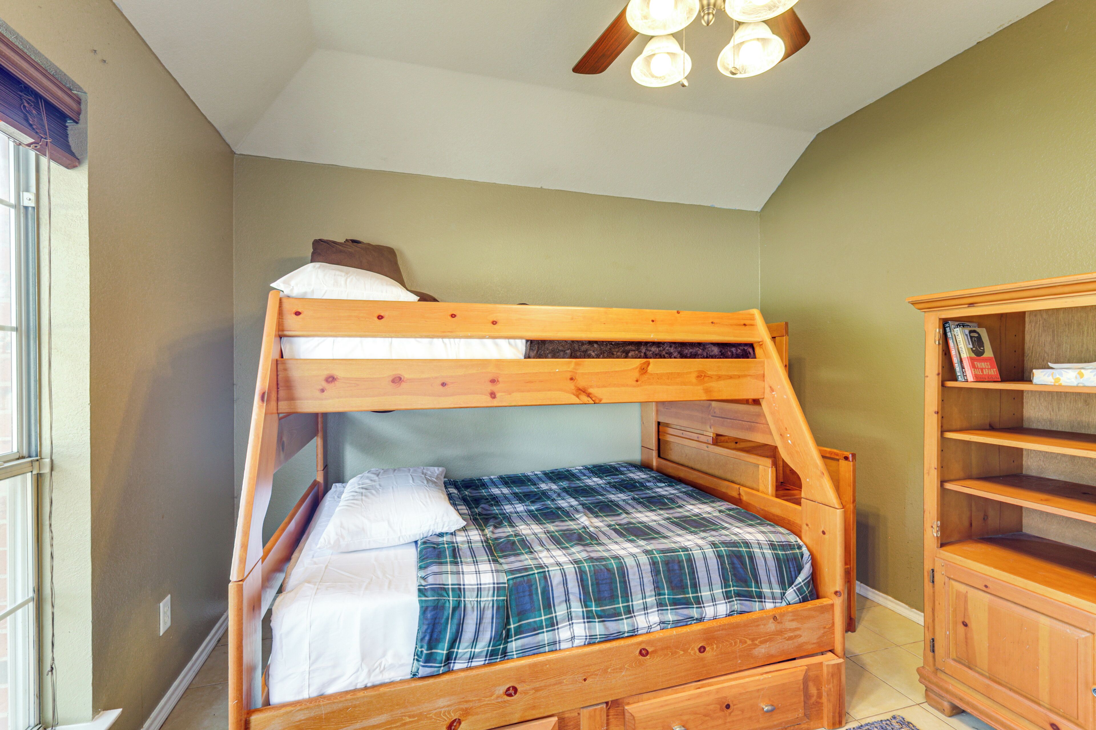 A bunk bed with a plaid bedspread and pillows, a wooden dresser with open shelves, and a ceiling fan with lights.