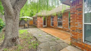 A brick house with a stone patio, a covered outdoor seating area, and a large tree in the foreground. - DFW Fun Awaits: Family Home in Crowley! (Crowley)