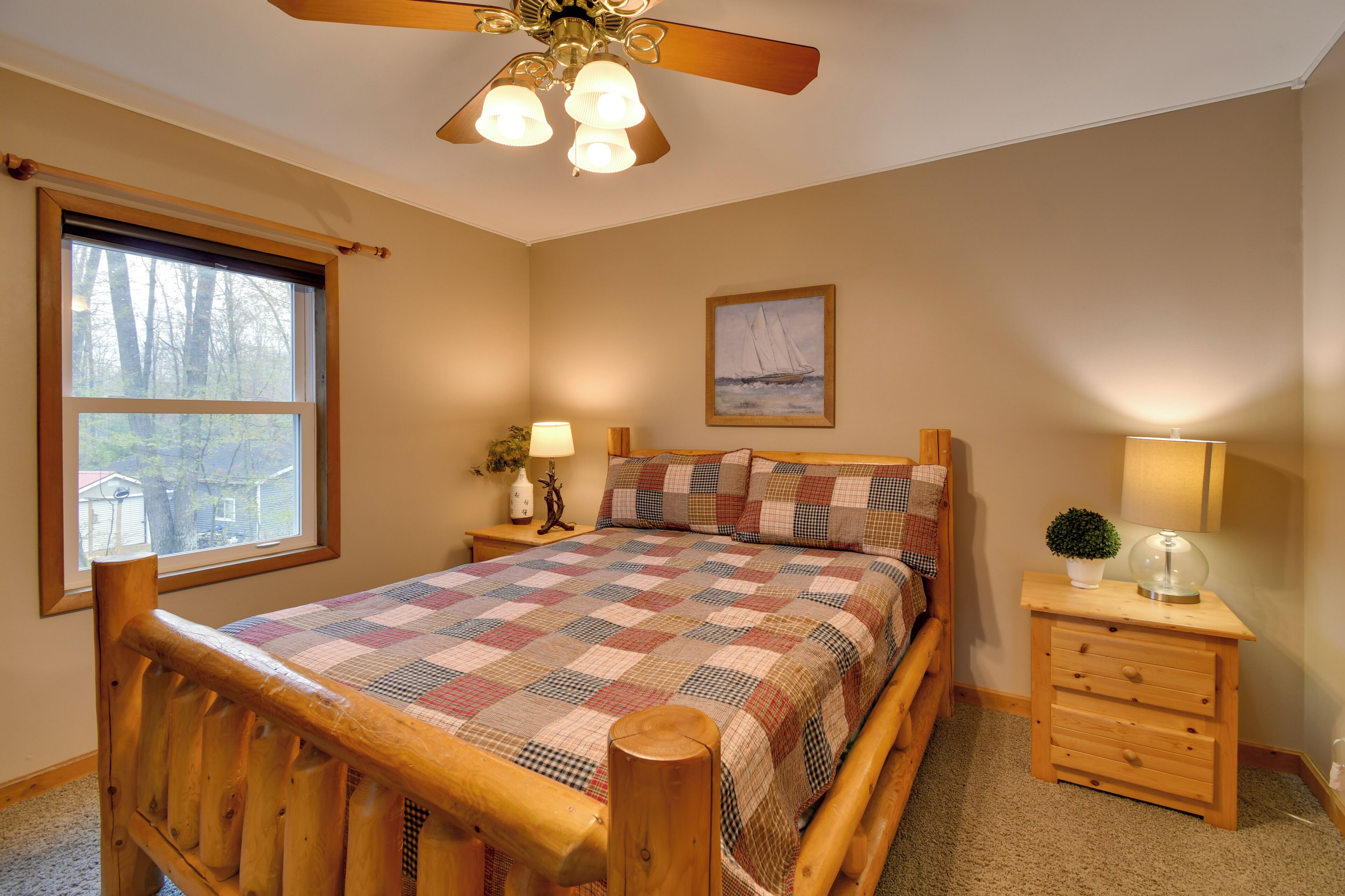 A bedroom with a wooden bed, a ceiling fan, a window with a view, a bedside table with a lamp, and a framed picture on the wall.