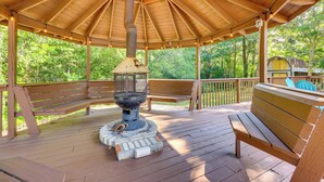 Une terrasse en bois couverte, dotée d’un foyer central, de bancs et offrant une vue sur la verdure environnante.