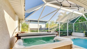 A hot tub under a covered patio with a view of a swimming pool and greenery.