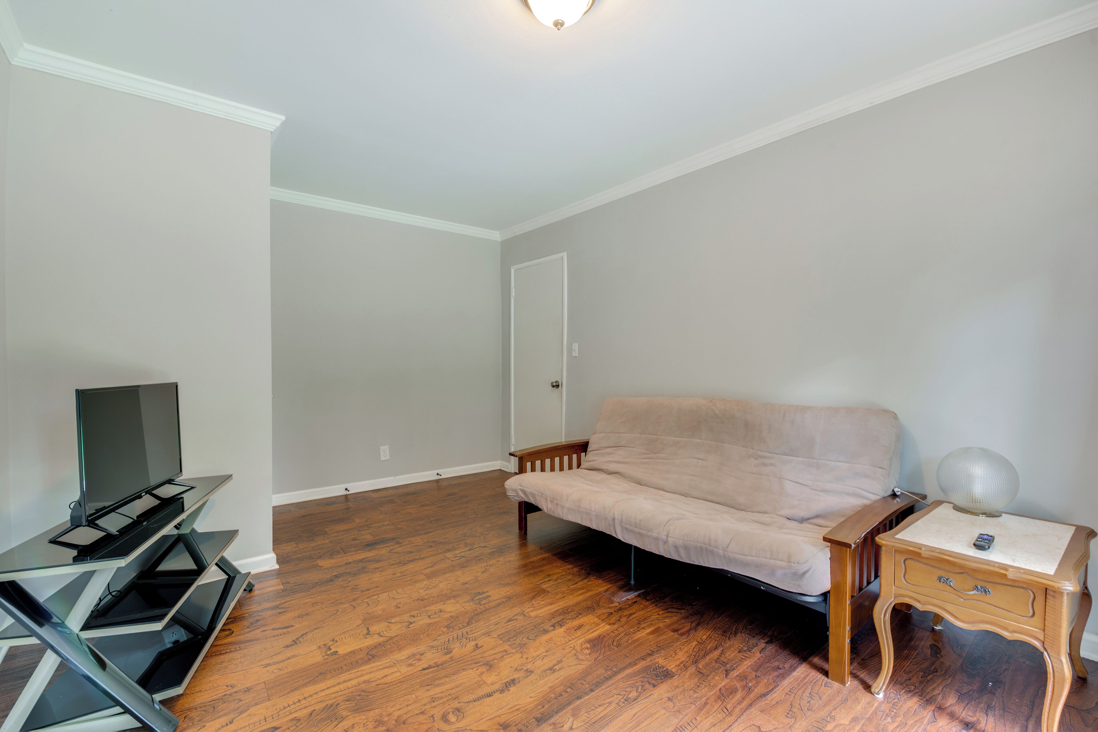 A room with a wooden floor, a beige sofa, a TV on a stand, a lamp on a nightstand, and a door.