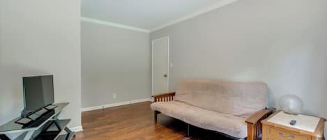 Quarto com piso de madeira, um sofá bege, uma TV em um móvel, uma luminária em uma mesa de cabeceira e uma porta.