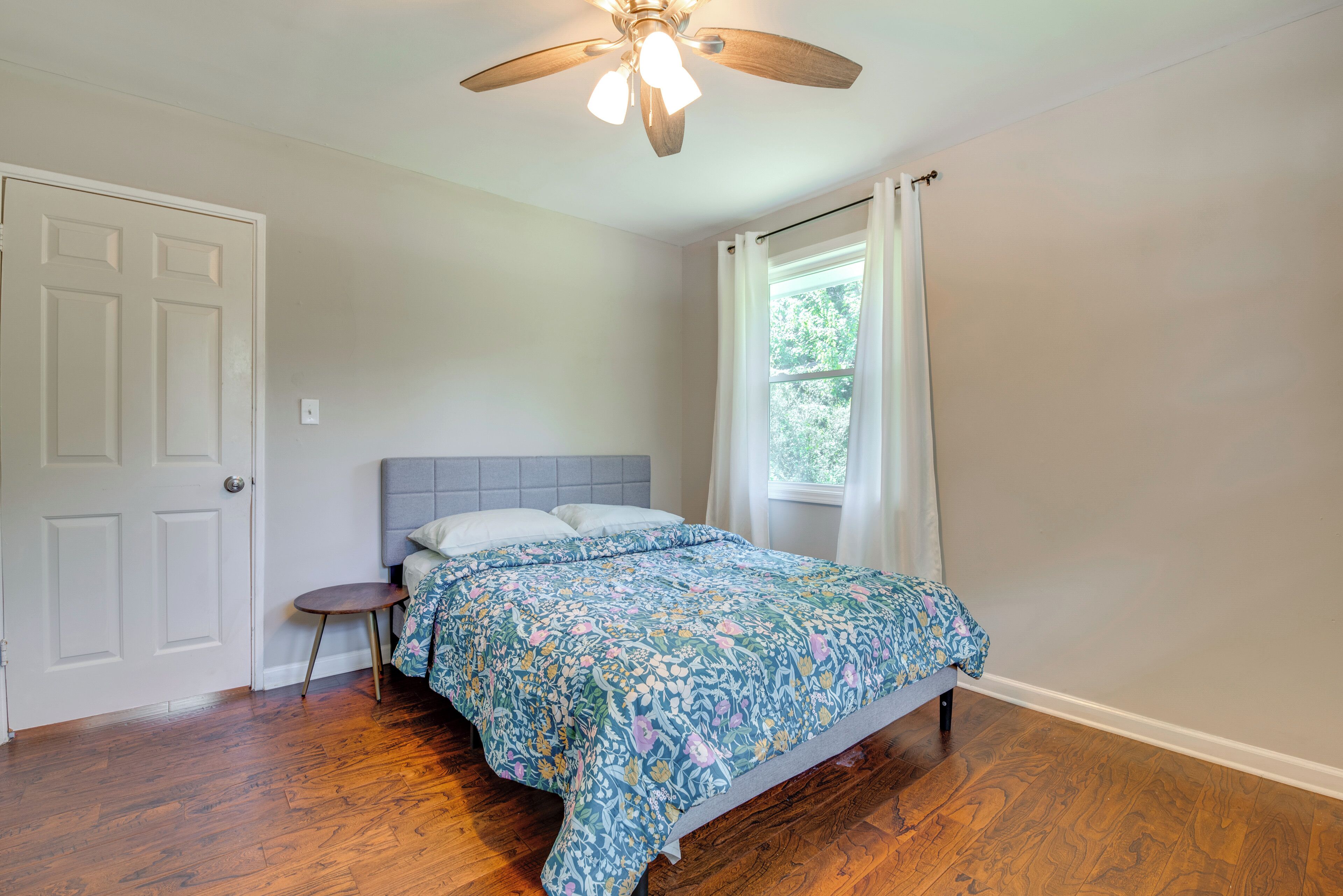 Une chambre à coucher comprenant un lit, un ventilateur de plafond, une fenêtre avec des rideaux et un plancher en bois franc.