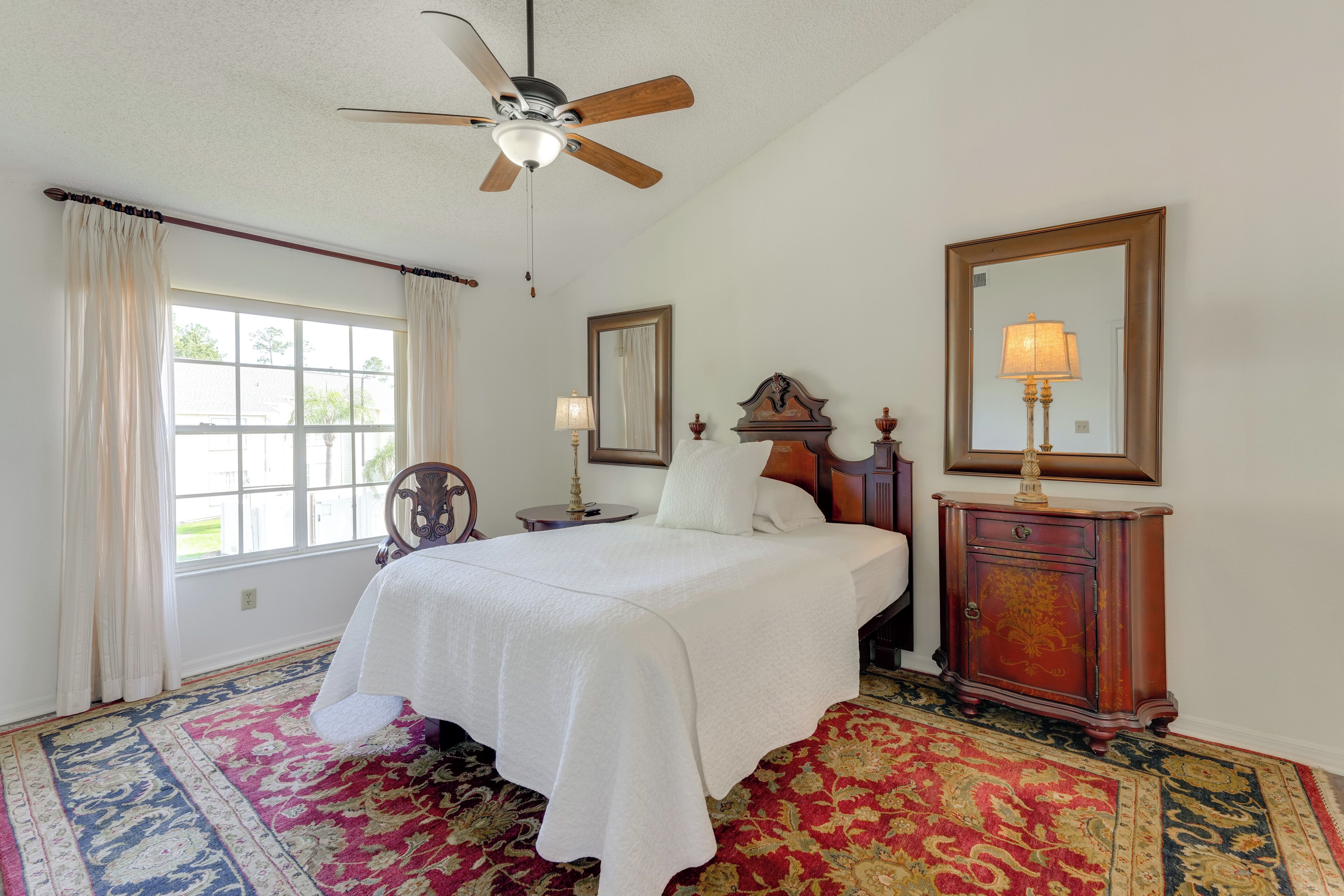 Une chambre avec un grand lit, un ventilateur de plafond, une table de chevet avec une lampe, une commode et une fenêtre avec des rideaux.