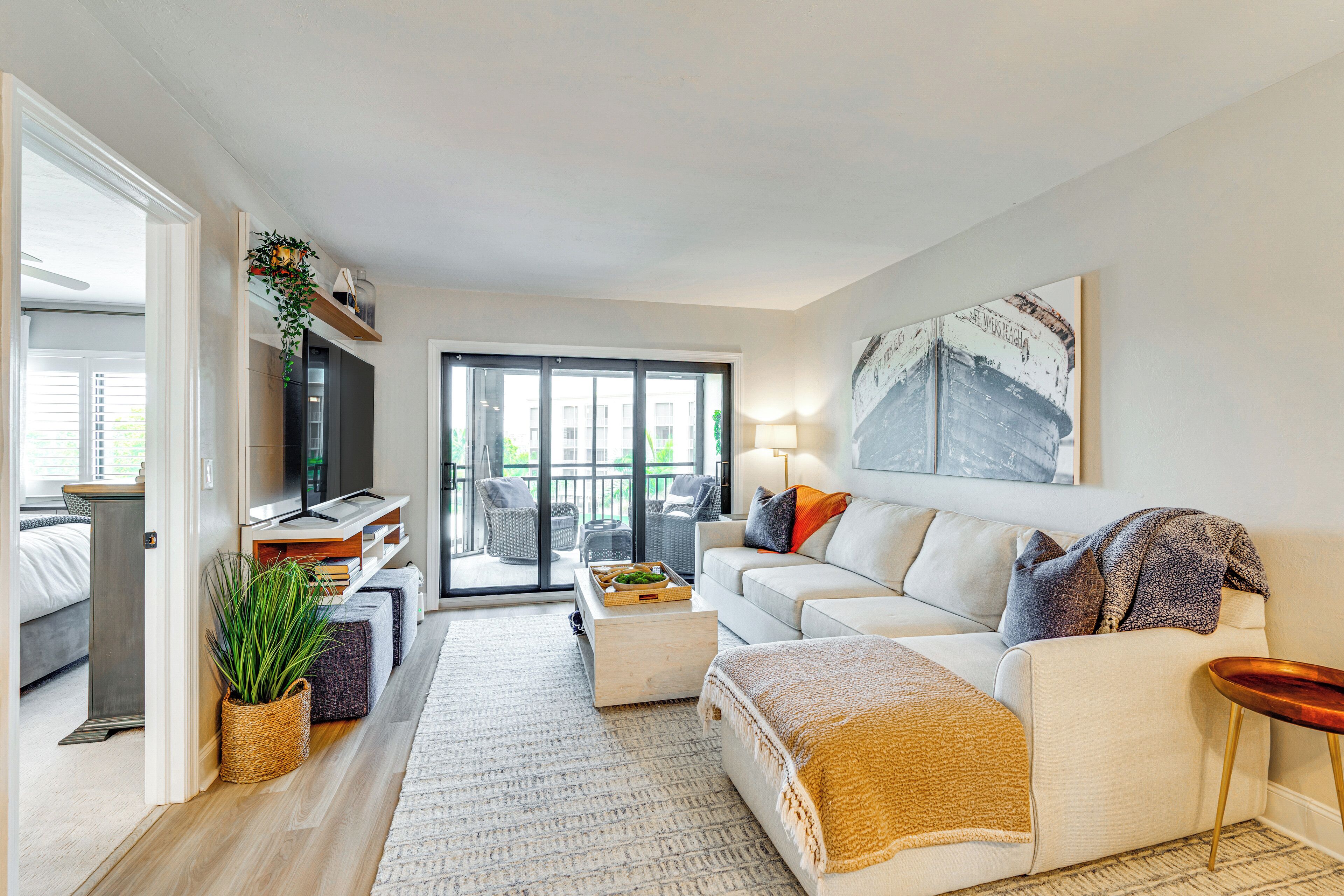 A modern living room with a sofa, coffee table, and a TV. There is a balcony with a view of buildings.