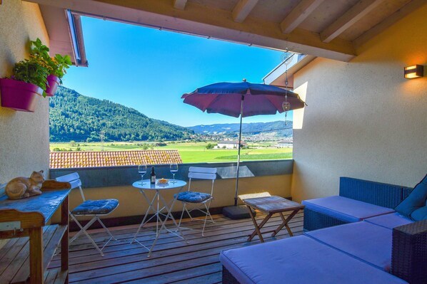 A balcony with a table, chairs, and an umbrella, overlooking a green landscape and hills.