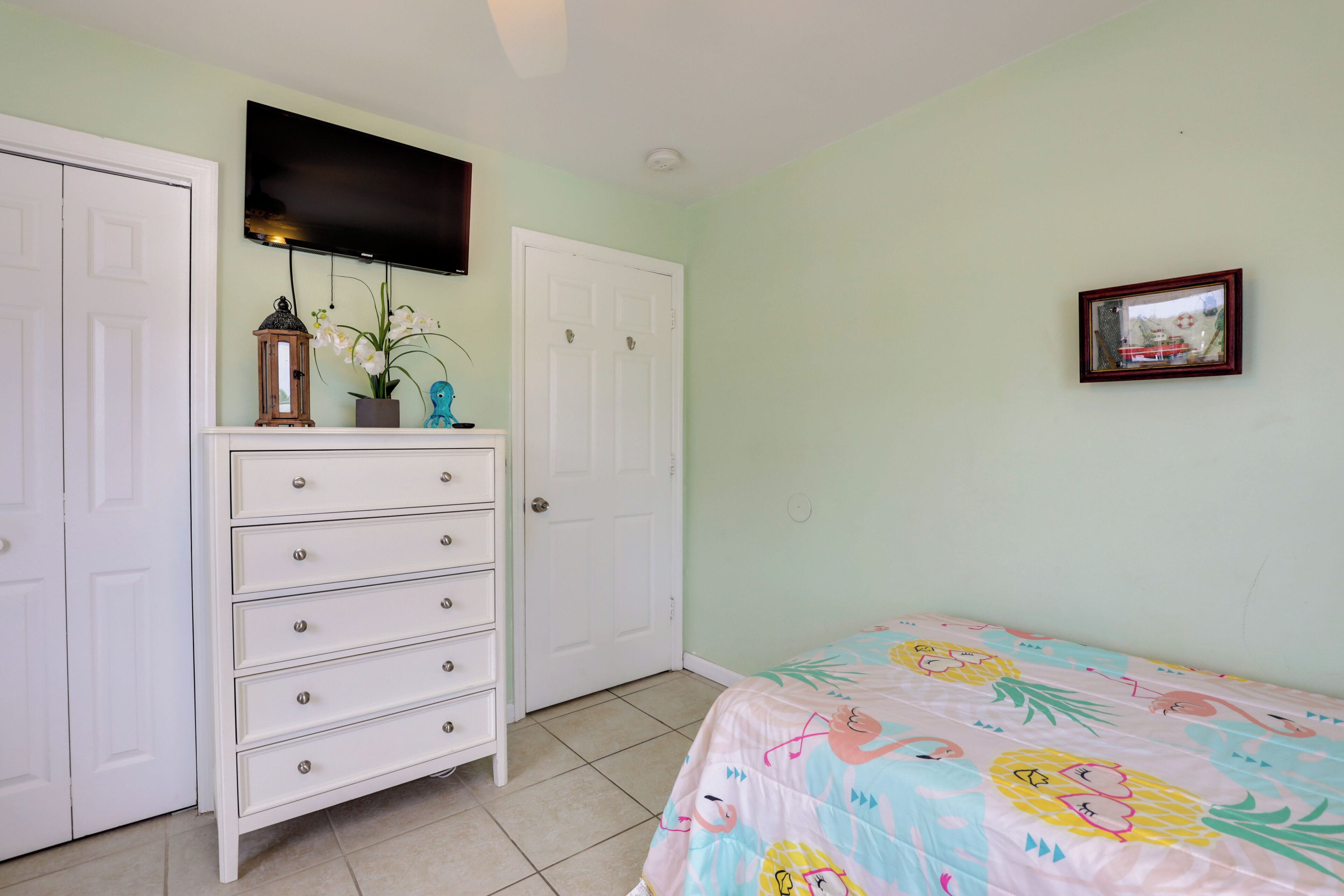 Quarto com cama, cômoda, televisão na parede e uma moldura com imagem.
