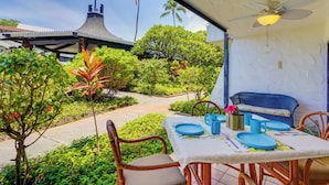 An outdoor dining area with a table set for two, surrounded by lush greenery and a view of a building with a distinctive roof. - 50 Feet to Ocean: Kona Deluxe Ground-floor Unit! (Kailua-Kona)