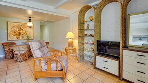 A living room with wicker furniture, a TV, and a decorative shelf. - 50 Feet to Ocean: Kona Deluxe Ground-floor Unit! (Kailua-Kona)