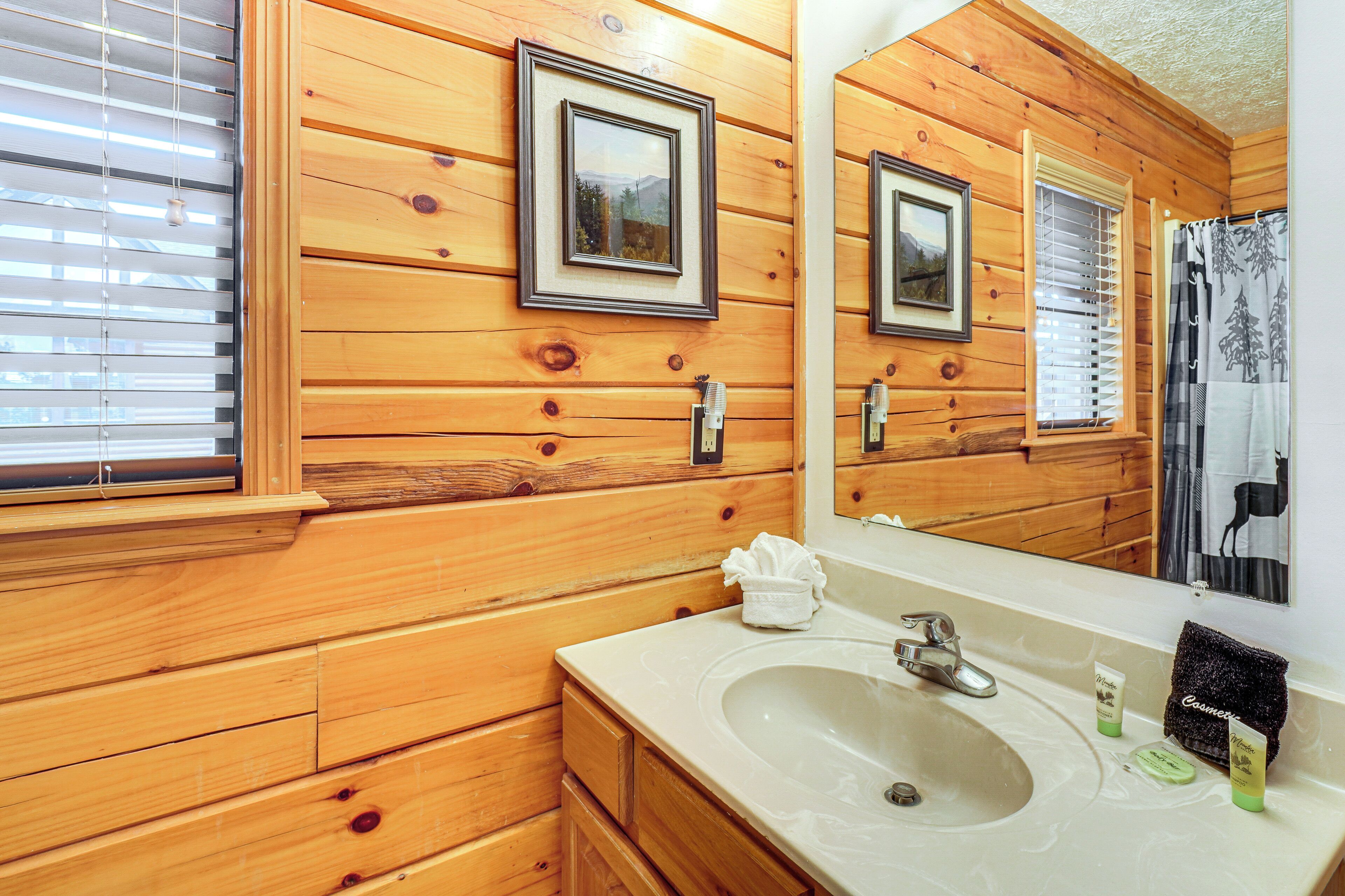 Une salle de bain rustique, avec des murs et un plancher en bois, un grand miroir, un lavabo et une fenêtre équipée de stores.