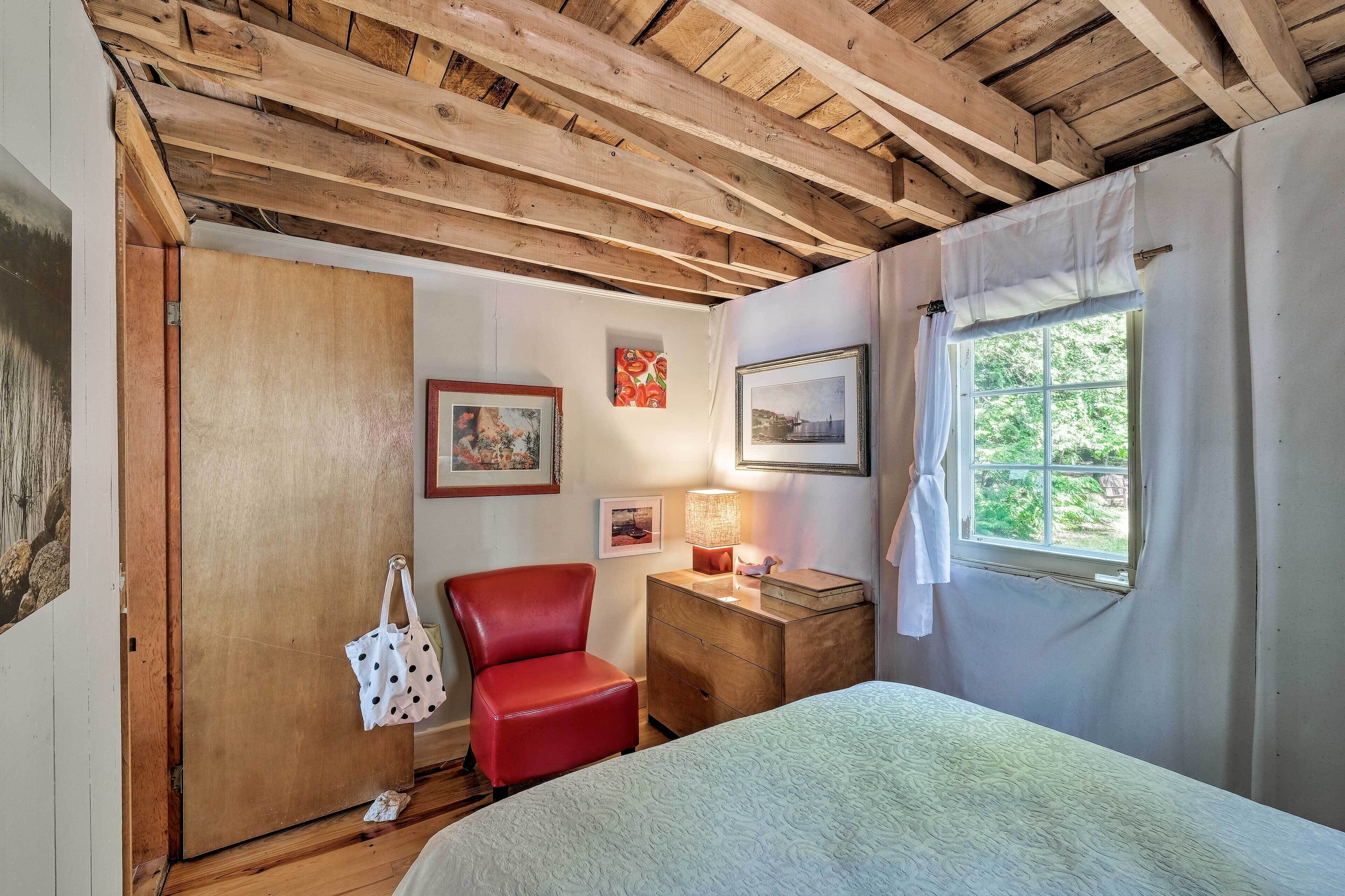 A bedroom with a wooden ceiling, a bed, a red chair, a wooden nightstand with a lamp, and a window with curtains.