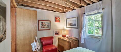 A bedroom with a wooden ceiling, a bed, a red chair, a wooden nightstand with a lamp, and a window with curtains.