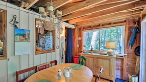 A rustic wooden cabin interior with a dining table, chairs, a chandelier, a window with a view of trees, and a calendar on the wall. - Cozy Lakefront Madison Cabin w/ Dock & Kayaks (Madison)